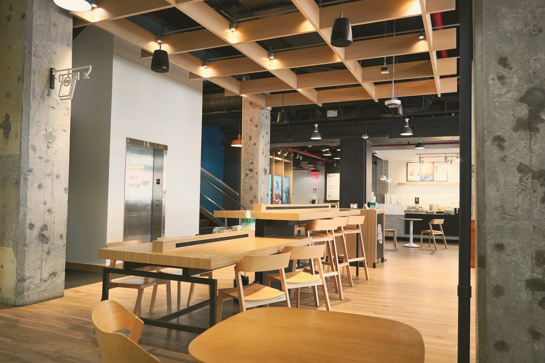Espace commun de bureau : tables et chaises en bois, plafond à caissons, poteaux en béton