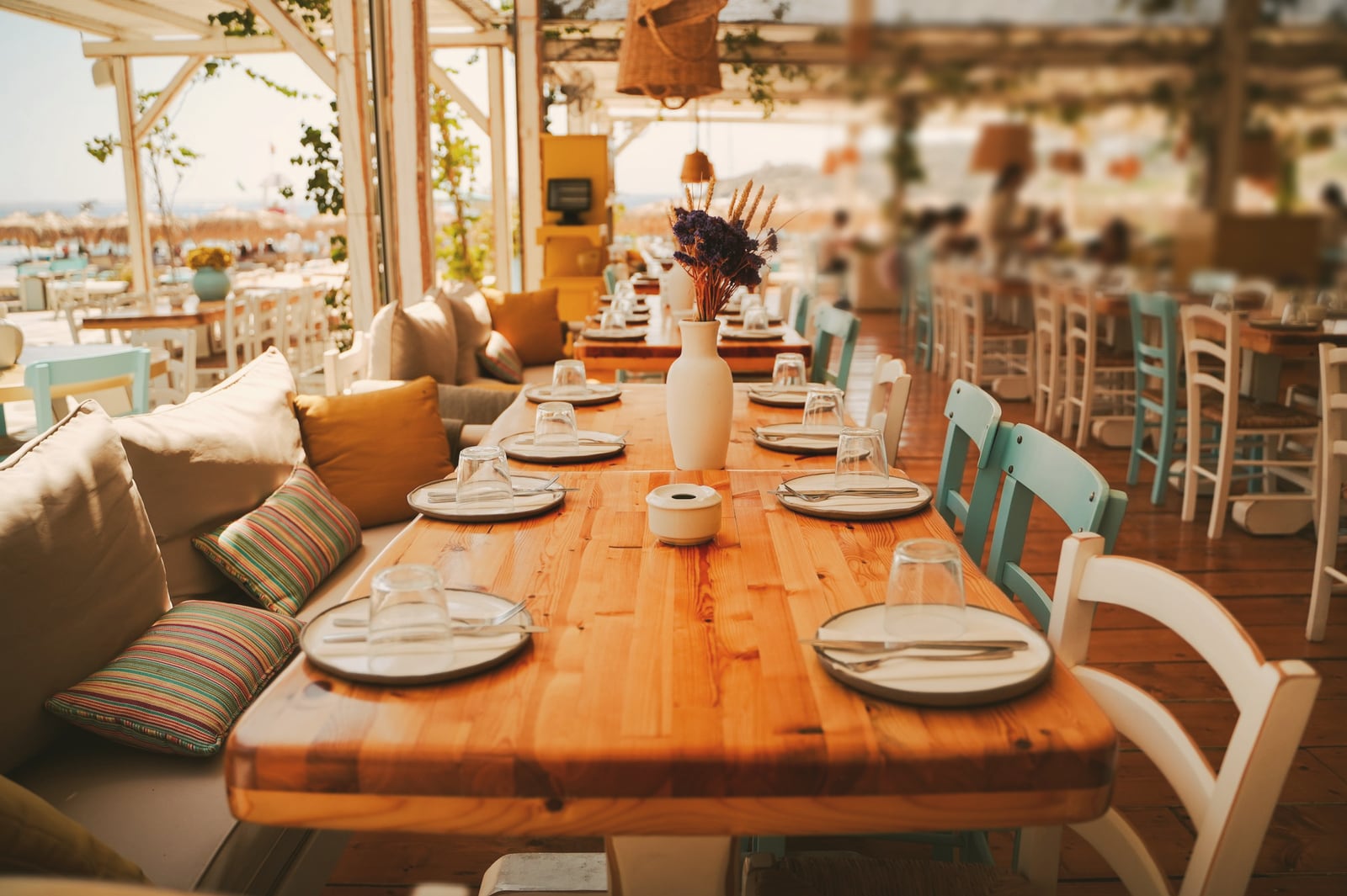 Terrasse de restaurant couverte : longue table en bois dressée, banquette à coussins et chaises en bois peint