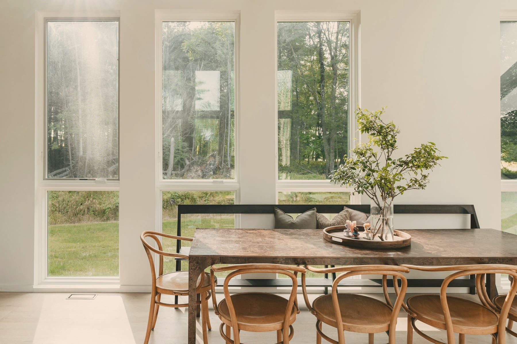 Salle à manger : table en bois sombre, banquette et chaises en bois courbé, grandes fenêtres sur la forêt